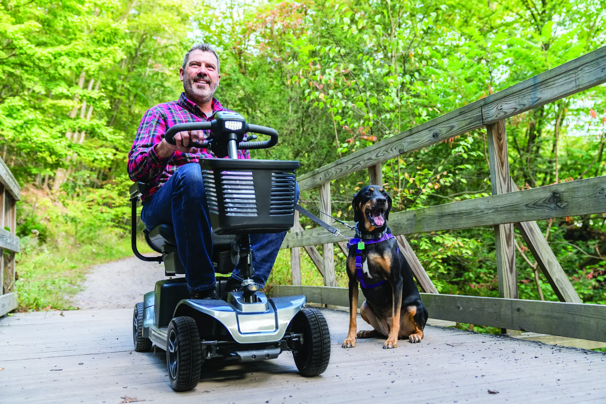 A man using a four-wheel mobility scooter on a wooden path with a dog beside him