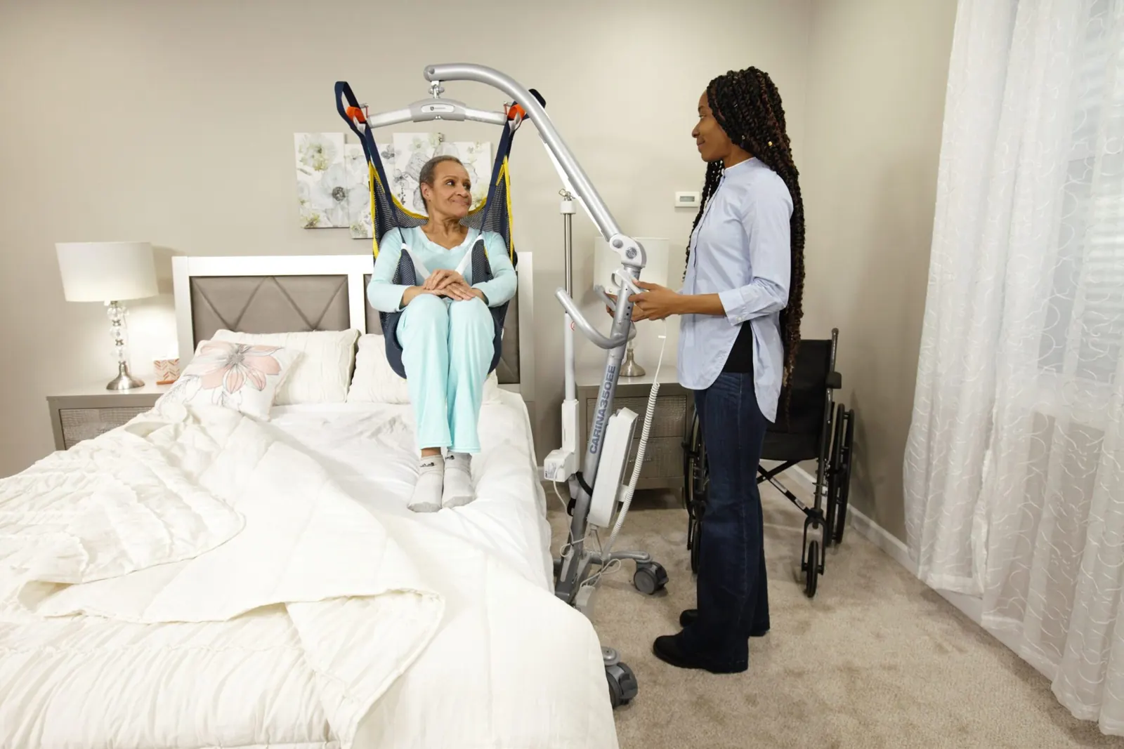 A caregiver beside a person supported in a full-body floor lift in a bedroom