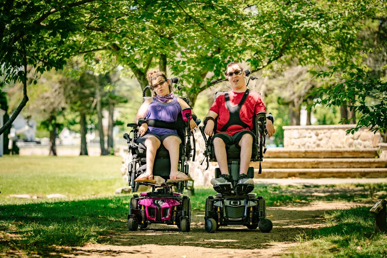 Two people using power wheelchairs together on a tree-lined park path