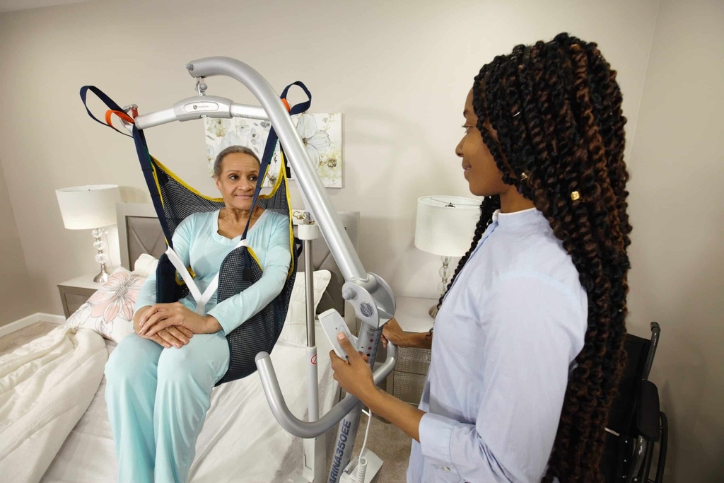 A person seated in a sling over a bed while a carer raises the Carina 350 lift