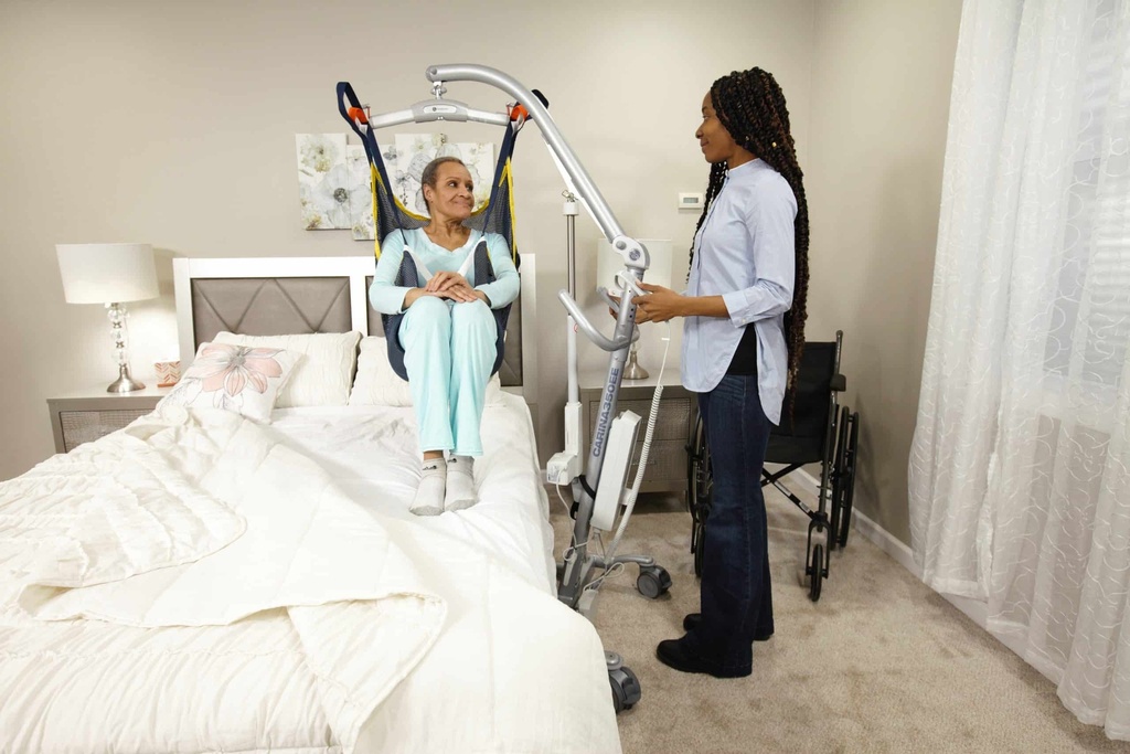 A person lifted clear of a bed in a sling on the Carina 350, with a wheelchair behind