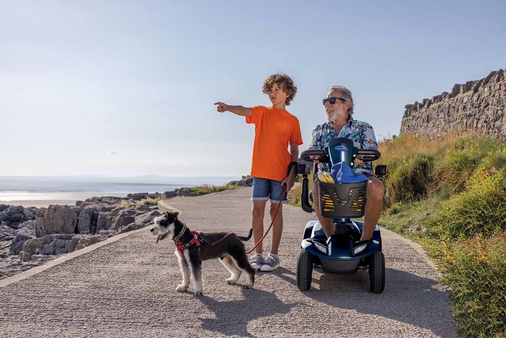 Man riding a blue Leo on a paved seaside path beside a boy walking his dog