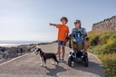 Man riding a blue Leo on a paved seaside path beside a boy walking his dog