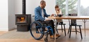 Person in a blue Helio Ultralight Folding wheelchair at a dining table beside a wood stove