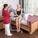 A resident seated on the Senta PUR L beside her care bed, with a carer at the hand control