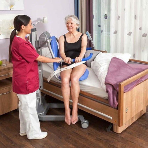 A resident seated on the Senta PUR L beside her care bed, with a carer at the hand control
