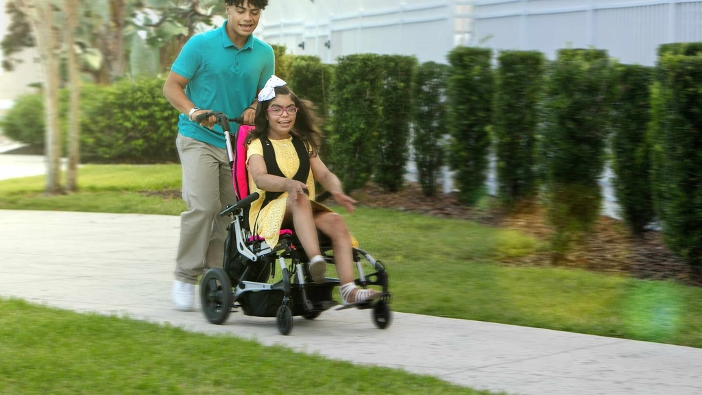 A caregiver pushing a child seated in a pink Sphynx along a paved path
