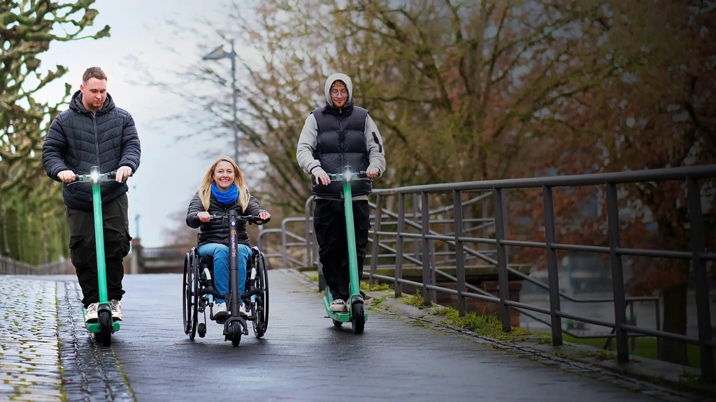 Rider steering the F35 on a damp path, beside two people on green electric scooters