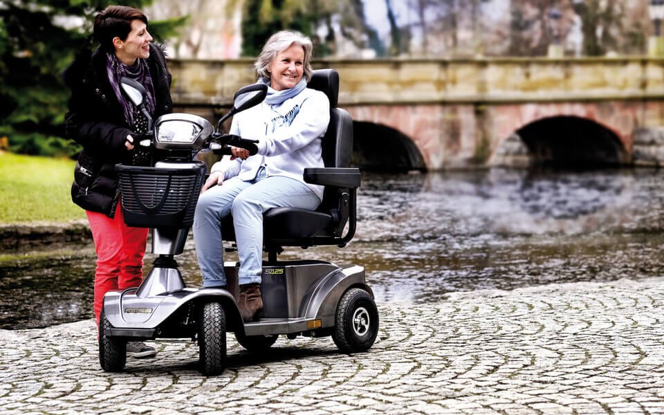 Fortress S425 mobility scooter in grey on a stone path, with a rider and a companion