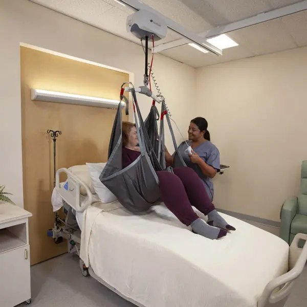Person in a grey sling over a bed, caregiver holding the lift hand control