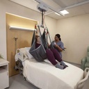 Person in a grey sling over a bed, caregiver holding the lift hand control