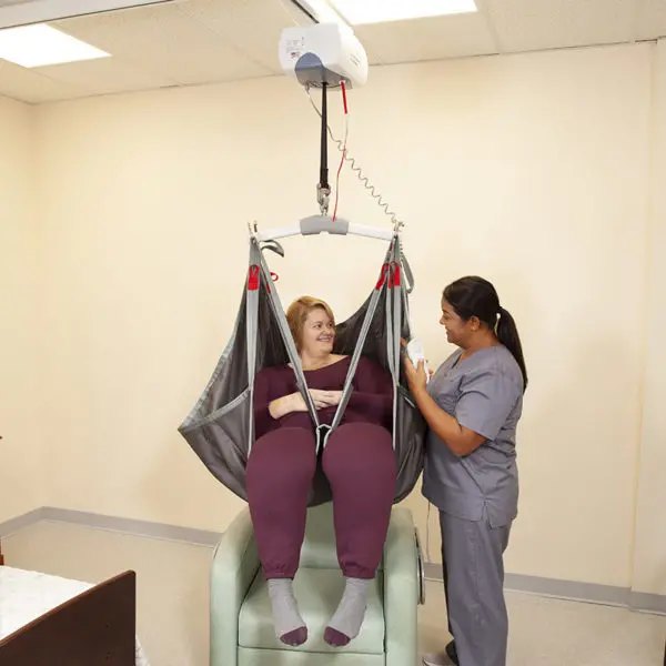 Two caregivers with a seated person in a grey sling under a ceiling track
