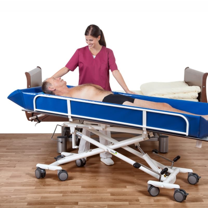 A resident lying on the Sina Comfort trolley beside a bed while a carer stands over him